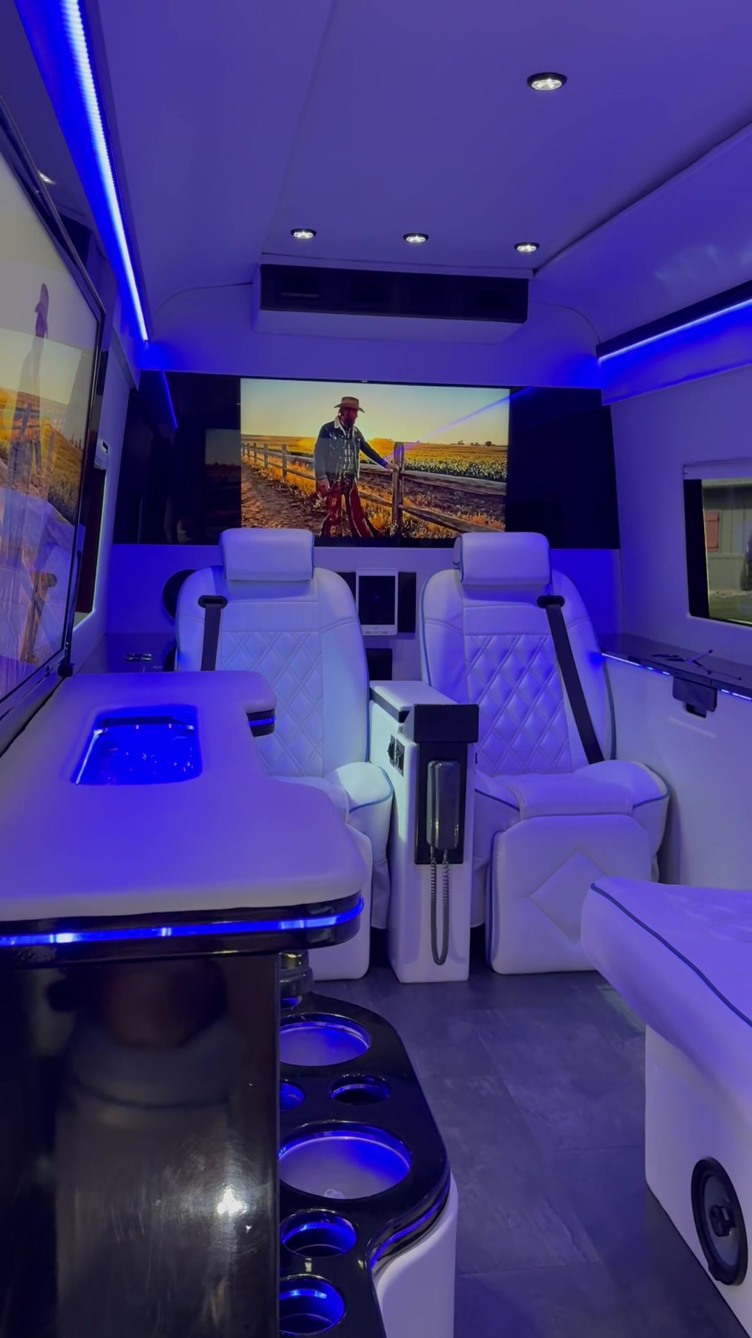 Interior of the luxury Mercedes Sprinter glowing in blue ambient light with white leather seating, a marble bar and flat-screen TVs