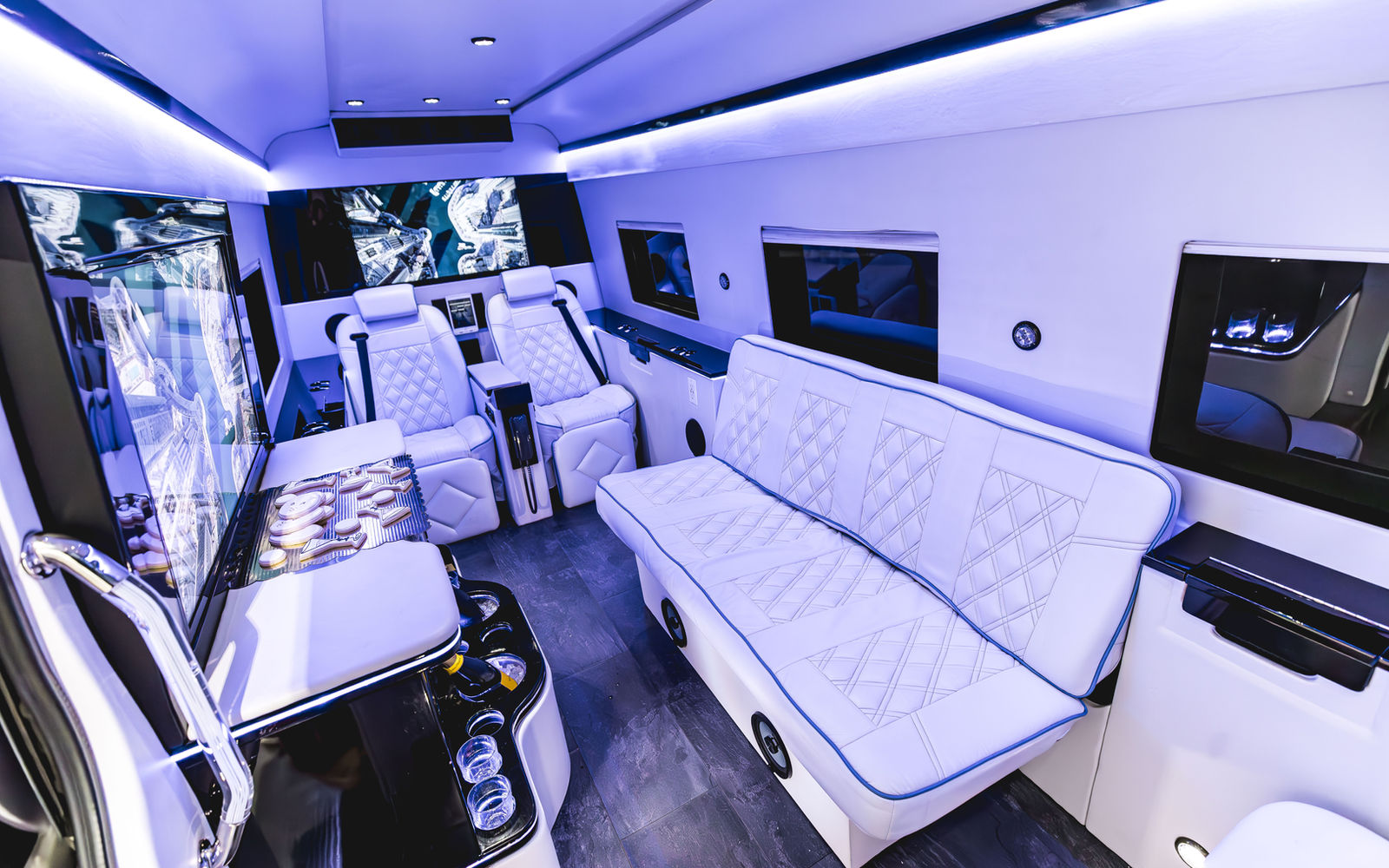 Interior of the luxury Mercedes Sprinter showing white captain's chairs, bench seating, bar and three flat-screen TVs